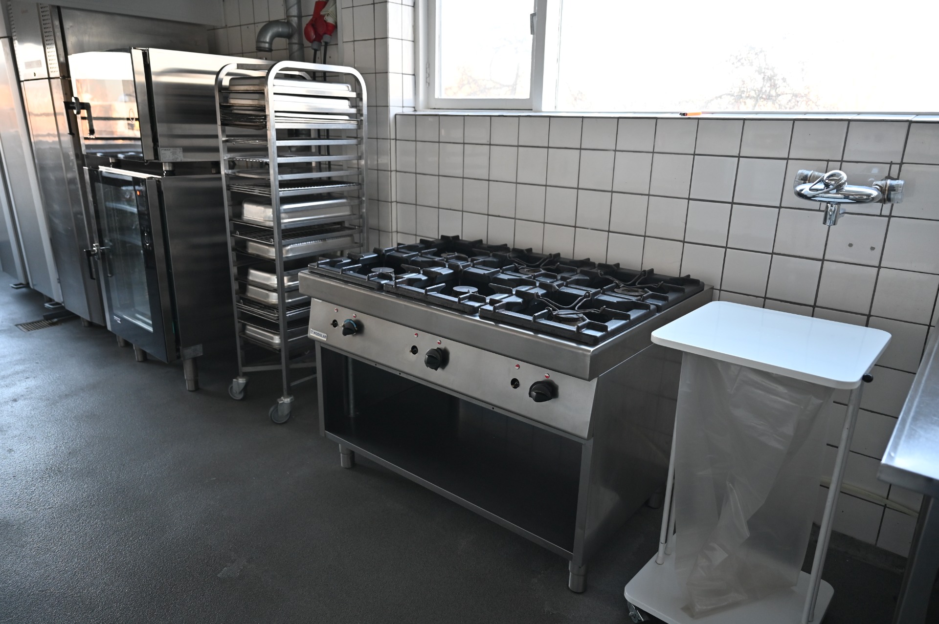 Rustfrit gasblusserbord med mange brændere i professionelt køkken, ved siden af ovn og vandhane.