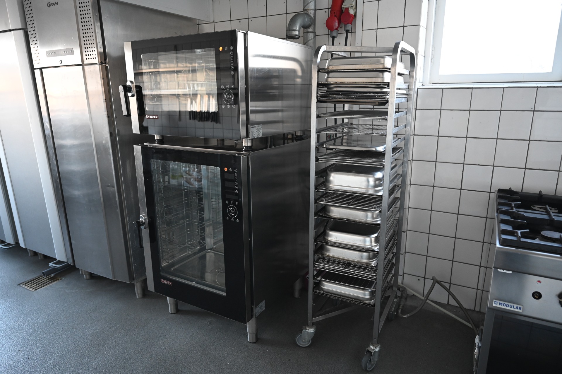 Rustfrit ovn- og dampudstyr med åbne døre, hylder og bakker i professionelt køkkenmiljø.