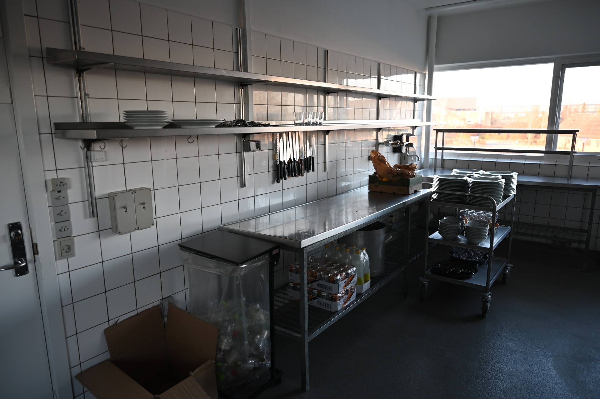 Professionelt køkken med rustfri arbejdsborde, opvaskestativer, stablede tallerkener og vindue mod udendørsområde.