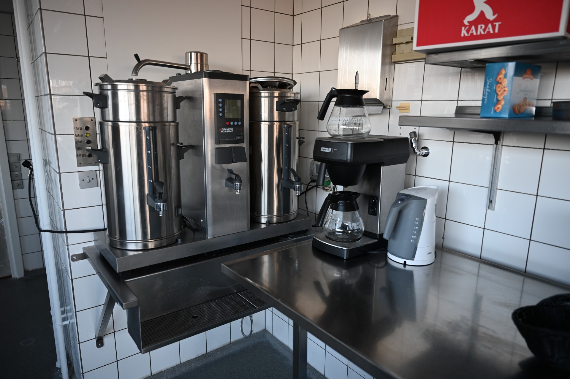 Rustfrit bryggeriudstyr med store kedler, kaffemaskine og glasbeholder på bord i flisebelagt rum.