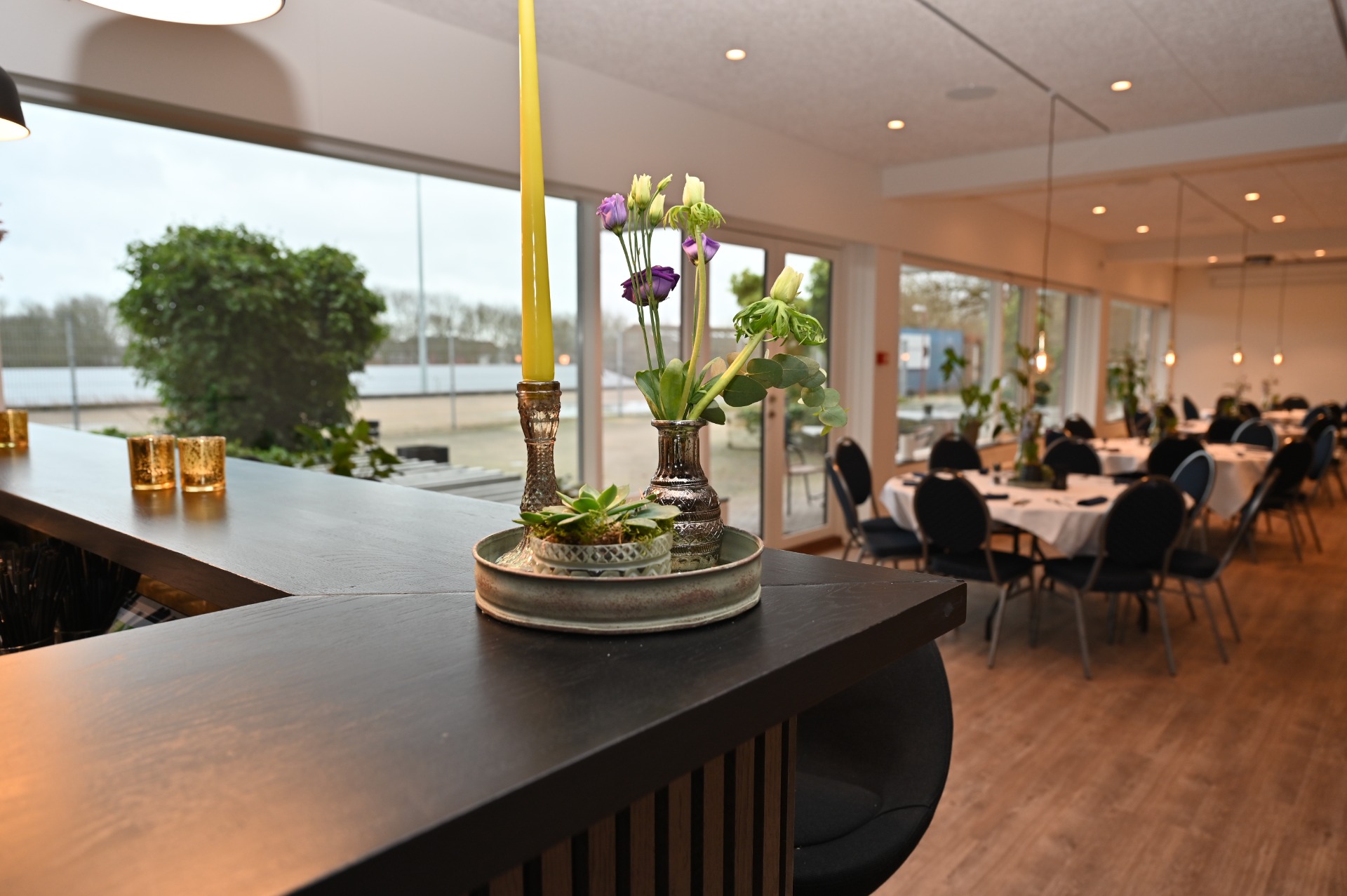 Blomsterarrangement i glasvaser på bord i lyst mødelokale med stole og vinduer i baggrunden.