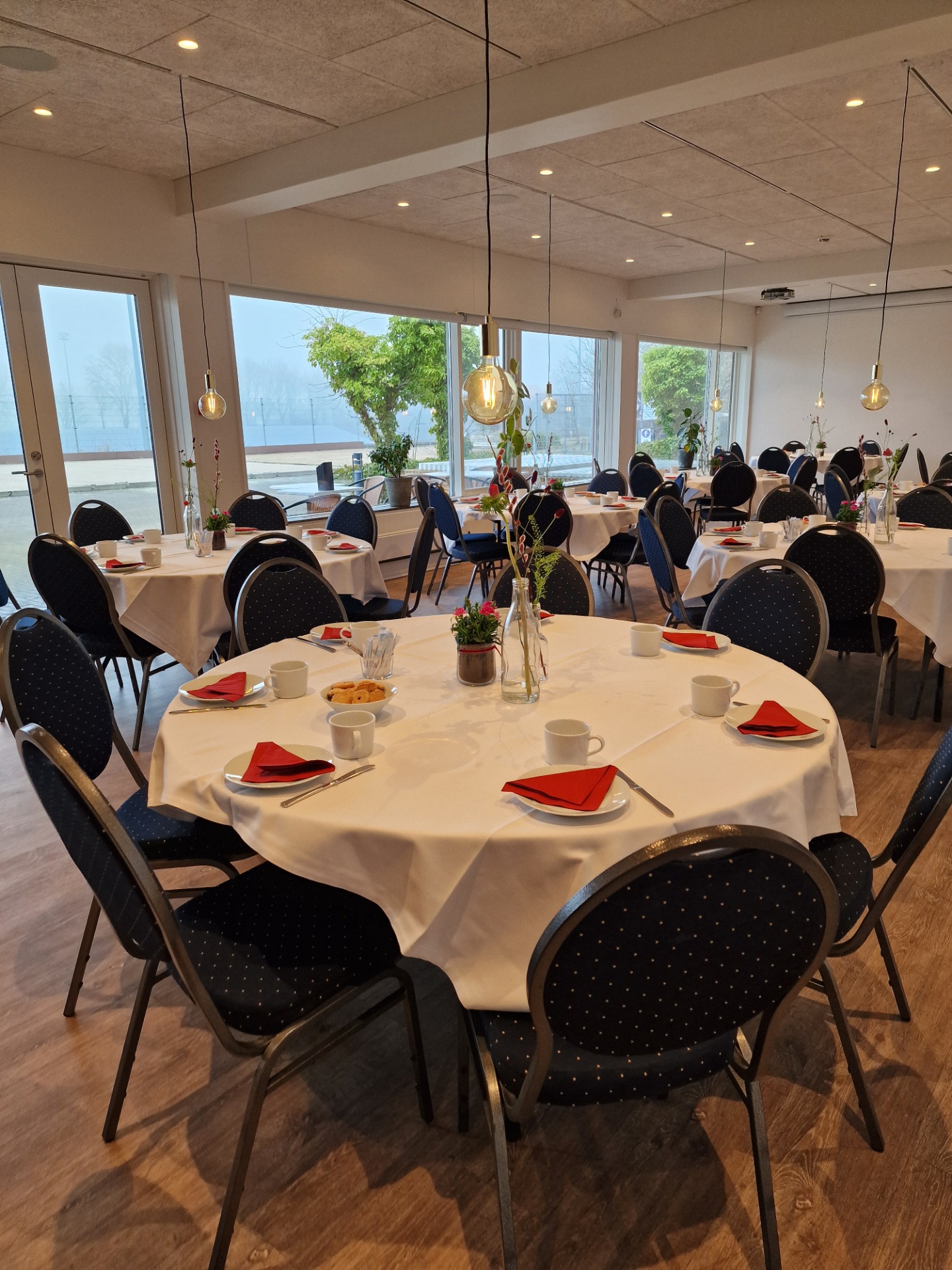 Dækket restaurantbord med hvide duge, røde servietter, sorte stole og hængende pærer i lyst lokale.